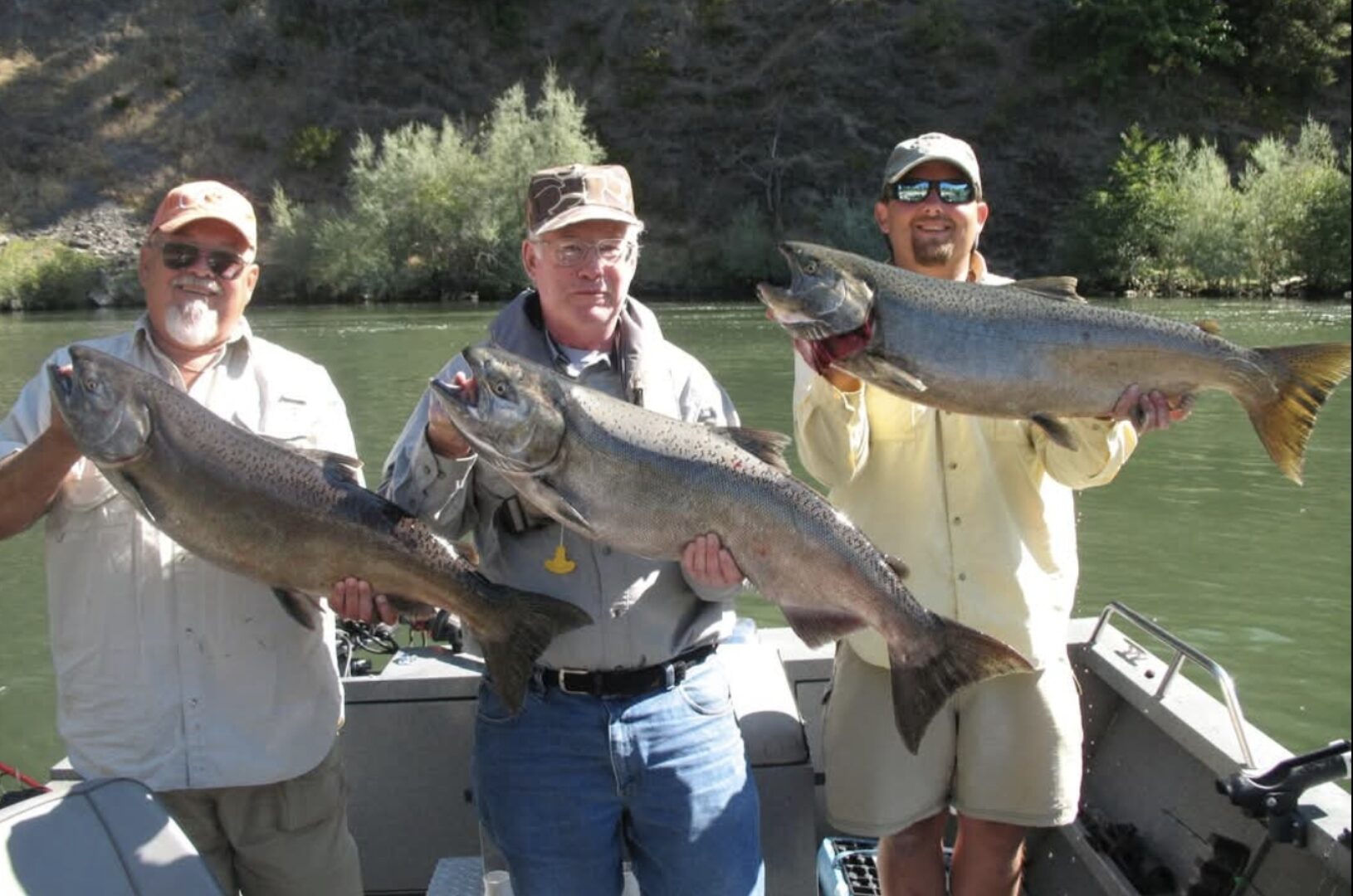 Three anglers holding large salmon on a fishing boat on a Southern Oregon river, with green hillsides and calm water in the background.