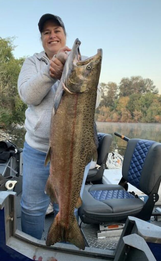Smiling angler holding a large salmon on a fishing boat in Southern Oregon, with calm river water and trees visible in the background.