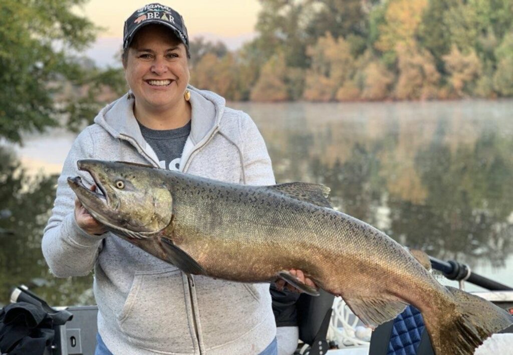 Smiling angler holding a large salmon on a calm Southern Oregon river, with fall-colored trees reflected in the water behind her.