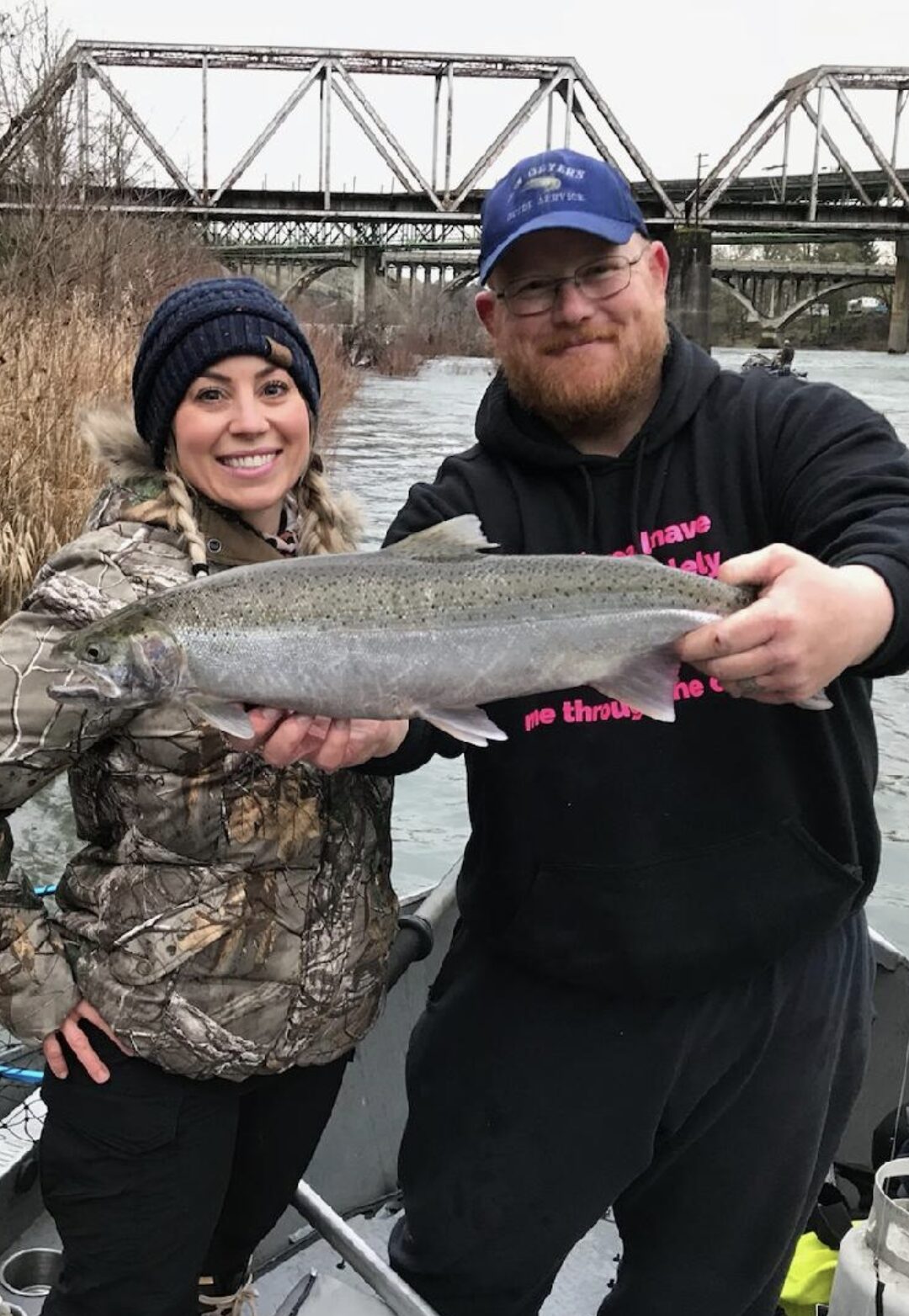 Two anglers holding a large steelhead on a boat on a Southern Oregon river, with a steel bridge and riverbank visible in the background.