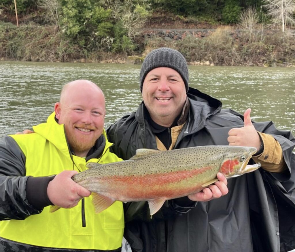 Two anglers smiling and holding a large rainbow trout on a Southern Oregon river, with flowing water and a wooded riverbank in the background.